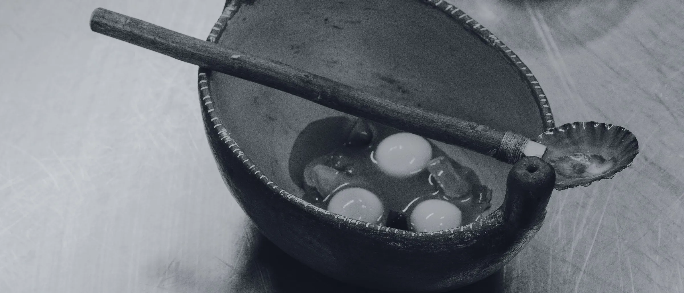 Potato spheres in broth served in a dark bowl, with a wooden pestle and a shell spoon, on the pass's steel counter
