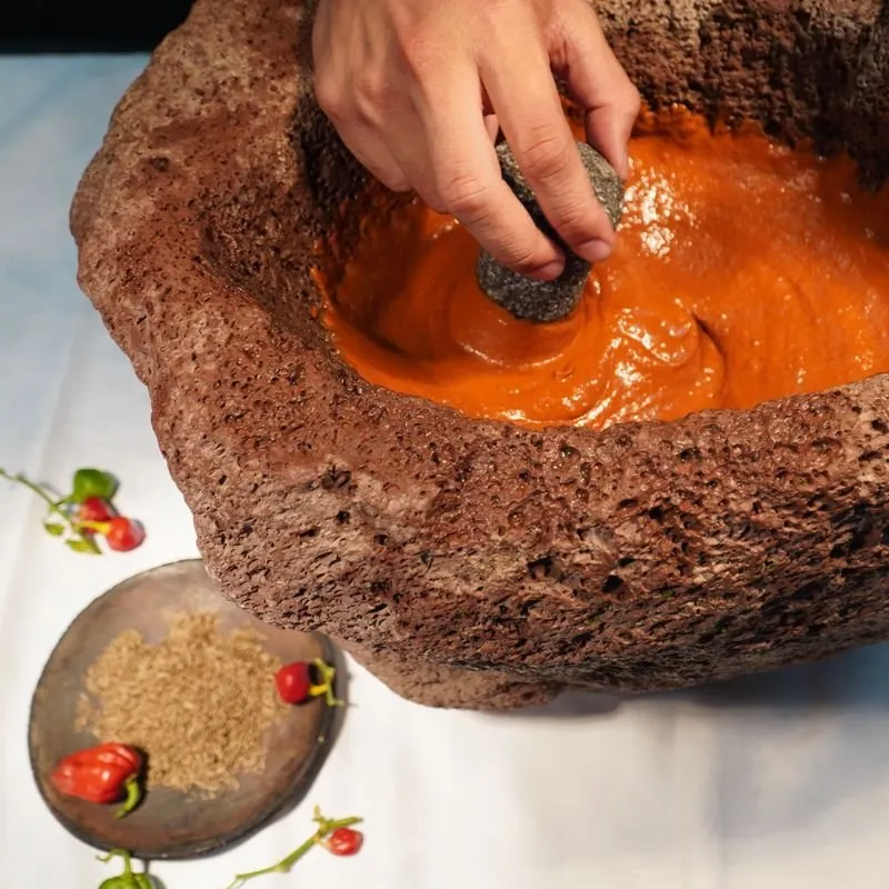 A hand grinding red mojo with a stone pestle in a volcanic stone mortar, chillies and cumin alongside