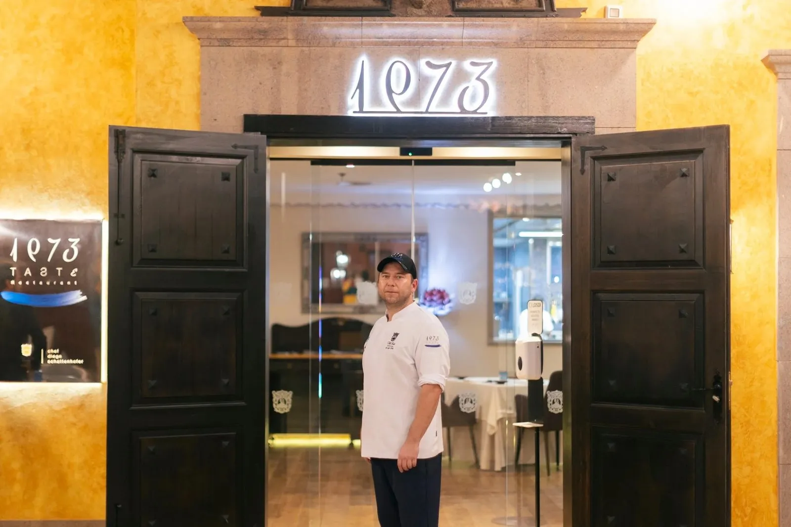 Diego Schattenhofer, in a white chef's jacket, at the entrance to Taste 1973 restaurant, under the illuminated '1973' sign
